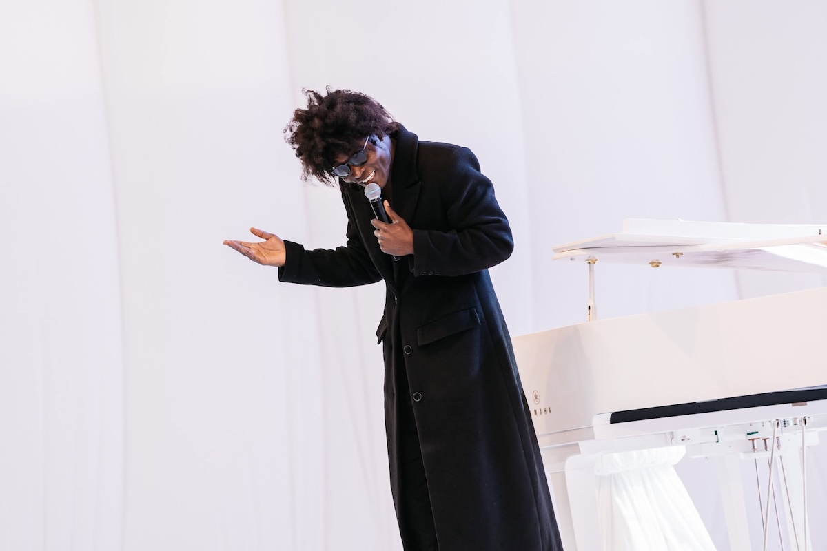 Musician Benjamin Clementine performs in a long black coat and sunglasses, holding a microphone next to a white grand piano.